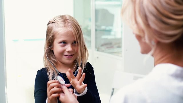 Cute kid girl with blond hair plays with stethoscope under supervision of doctor at pediatrician appointment. Little girl taps stethoscope with her hand and listens to the beats. Shot in slow motion