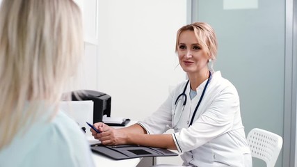 Young blonde woman at doctor reception in white dressing gown. Therapist female sitting at table and asks the patient questions. Shot in slow motion