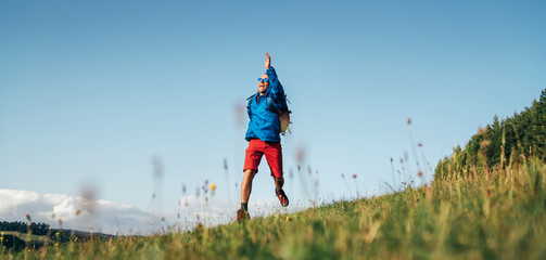 Backpacker traveler emotionally jumping over green grass mountain meadow with backpack with wide...