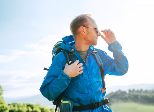 Backpacker man lifestyle side portrait enjoying mountain landscape. He wears in blue rain coat poncho and blue sunglasses. Active sports backpacking healthy lifestyle concept.