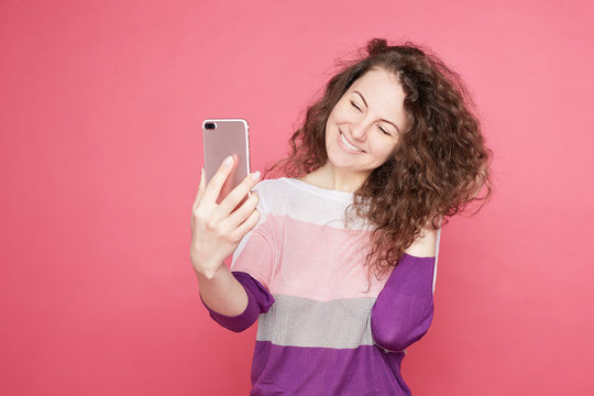 Lovely glad female smiles broadly, takes selfie portrait on cell phone, enjoys free time, makes pics for social networks, wears colorful blouse, posing on pink wall background, smiles at mobile camera