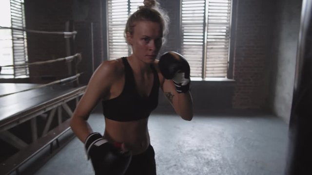 Medium shot of attractive muscular blonde sportswoman wearing boxing gloves punching boxing bag then stopping, wiping forehead with her hand and looking at camera