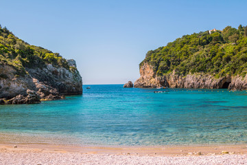 Fototapeta premium Seascape – lagoon with turquoise water, mountain with cliffs, green trees, blooming bushes, rocks in a blue water, colorful cruise touristic boats. Corfu Island, Greece. 