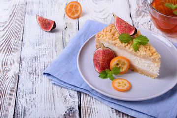 Piece of grated pie with cottage cheese topped with figs and kumquats on ceramic plate on white wooden table