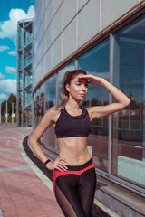 Fototapeta premium girl in summer in city, sportswear on run in morning closes her palm from bright sun, looks into distance. Search for friends girlfriends at a fitness workout , lifestyle healthy way of thinking.