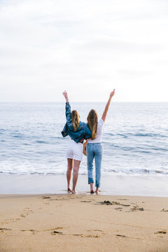 Two cool girls standing on sand at the beach with hands raised up