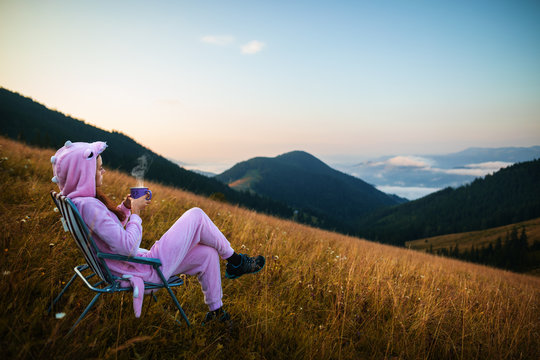 Young girl in a pink dragon costume drinking coffee while sitting in a chair on top of a mountain. Incredible mountain landscape at sunrise. Girl in pink pajamas drinks hot drink in the mountains