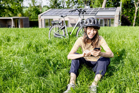 Laughing woman with bicycle using cell phone on a meadow in front of a house