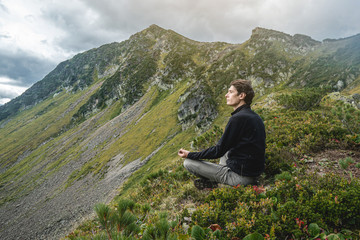 Fototapeta premium Man hiker sitting in a yoga pose at the peak of the mountain in the summer. Meditation after a long climb on a mountain