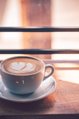 Latte coffee in white coffee cup on the wooden table in front of the large window of the coffee house.