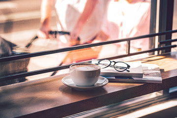 Latte coffee in white coffee cup on the wooden table in front of the large window of the coffee house.