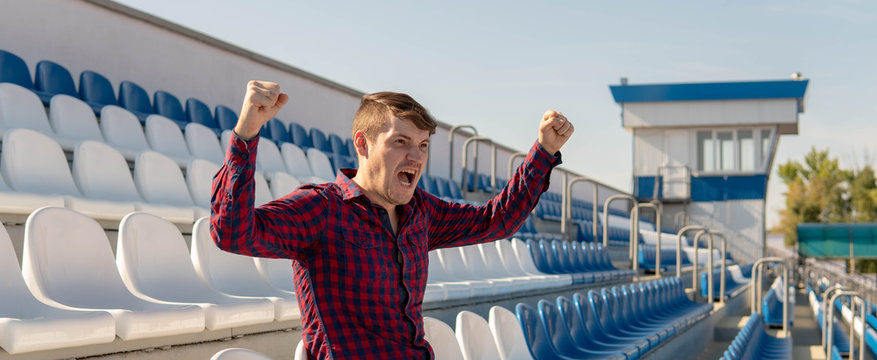 A young handsome man in a plaid shirt is supporting and rooting for his a favorite sports team on a stadium bleachers alone.