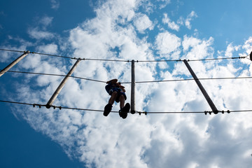 Boy climbing rope trail viewed from bottom in an adventure park. Silhouettes in front of a cloudy sky background.