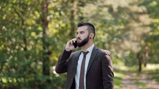 Young attractive male businessman in a brown suit walks in a green summer park and talk on phone. Smiling in a positive mood.