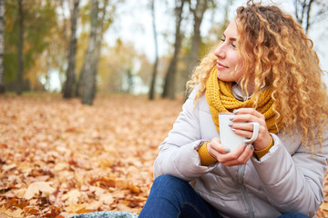 Redhead curly cheerful cute girl siting in autumn park, and holding coffee to go.