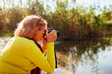 Middle-aged woman checking images on camerasitting by autumn river bank. Senior woman enjoying hobby