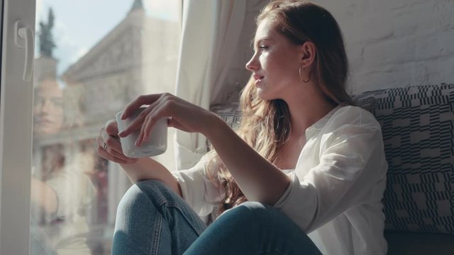 Close-up portrait of charming young woman with frizzy hair sitting in the room enjoying a cup of tea and looking through the window at the city