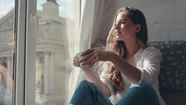 Gorgeous young woman in the house holding and drinking a cup of tea near the window smiling looking outside feel happy