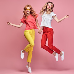 Two fashionable girl jump Smiling in colorful outfit on coral. Beautiful easy-going woman in red yellow pants, Stylish curly hair having fun. Joyful funny slim sisters friends, happy fashion concept