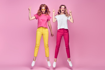 Two fashionable girl jump Smiling in colorful outfit on pink. Beautiful easy-going woman in red yellow pants, Stylish curly hair having fun. Joyful funny slim sisters friends, happy fashion concept