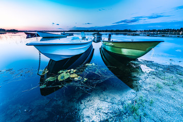 Motor and row boats anchored on the calm summer lake, sunset time, boats reflected in the water. Stocksjo Lake, close to Umea city, Northern Sweden