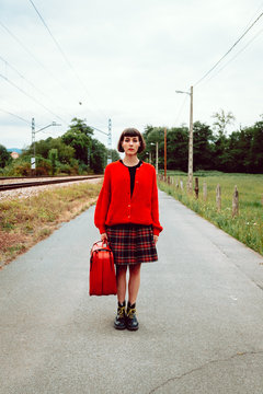 Woman in stylish boots with red suitcase standing on road