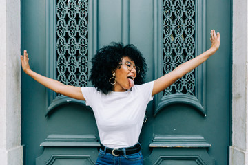 Young African American woman standing with closed eyes by green wooden door and sticking out tongue on daytime