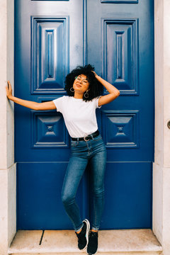 Pretty ethnic woman in white t-shirt and jeans leaning on blue door and looking up with closed eyes