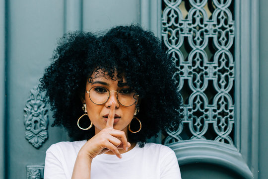 Pretty ethnic woman in white t-shirt and jeans leaning on green door and looking at camera with finger on lips like silence sign