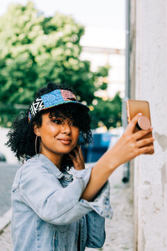 Beautiful African American woman in denim jacket and colorful cap visor standing with smartphone and taking photo in city street
