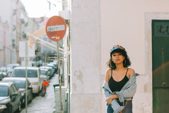 Fashionable African American woman in tank top and unbuttoned denim jacket standing on sidewalk and looking at camera
