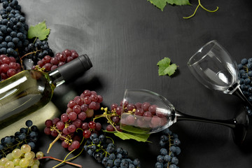 White wine bottle, two wineglasses, fresh grapes and leaves on black wood background. Flat lay, top...
