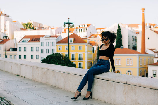 Young trendy African American woman in jeans and crop top sitting on stone parapet and looking over sunglasses