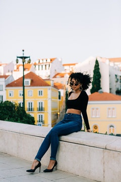 Young trendy African American woman in jeans and crop top sitting on stone parapet and looking over sunglasses