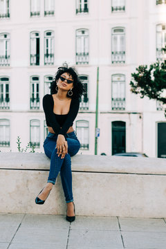 Young trendy African American woman in jeans and crop top sitting on stone parapet and looking over sunglasses