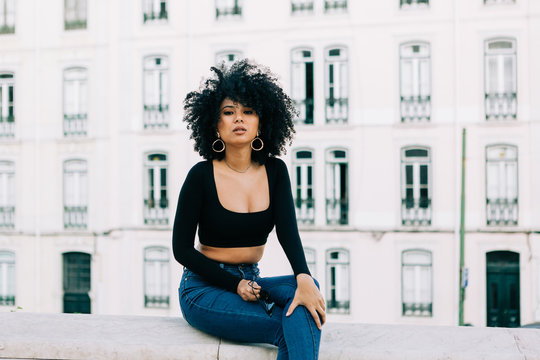 Young trendy African American woman in jeans and crop top sitting on stone parapet and looking at camera