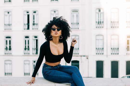 Young trendy African American woman in jeans and crop top sitting on stone parapet and looking over sunglasses