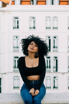 Young trendy African American woman in jeans and crop top sitting on stone parapet and looking at camera