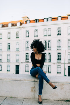 Young trendy African American woman in jeans and crop top sitting on stone parapet