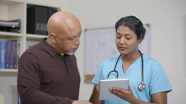 Nurse w/ stethoscope reviews info on tablet at nursing station, camera move left