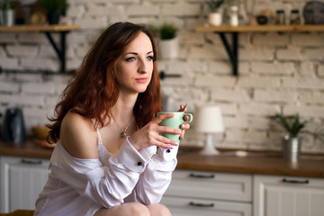 Adorable nice charming cheerful pretty trendy stylish girl drinking coffee, sitting on table countertop in modern light white kitchen