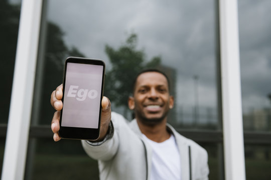 Stylish cheerful African American man holding smartphone with Ego text on screen on background of reflecting wall