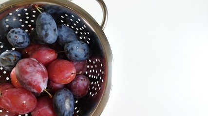 A fragment of a metal colander with Red and blue plums on a white background. Water droplets and holes in the metal. Macrophoto, Food photo, flat layout, top view. copy space for text. The subject is