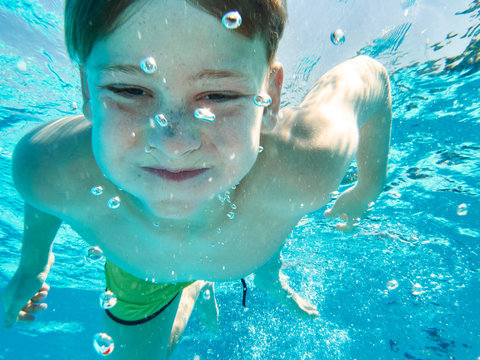 Squinting boy in shorts swimming under water of pool holding breath and looking at camera