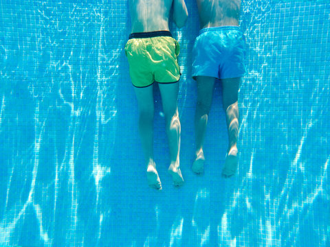 Crop back view of two boys in swimming shorts floating underwater in clear water of pool