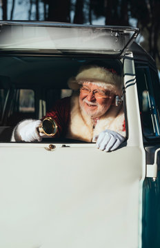 Smiling senior man in costume of Santa Claus driving old van with metal bell in hand on sunny day looking away