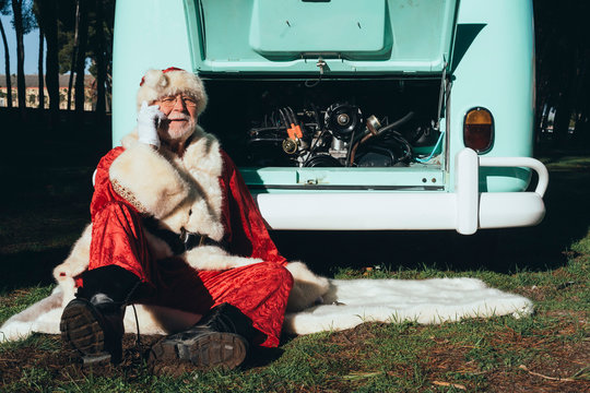 Senior man in costume of Santa Claus speaking on mobile phone while sitting by van with open engine compartment