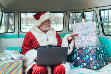 Cheerful man in costume of Santa Claus sitting in van with laptop and colorful gifts on sunny day