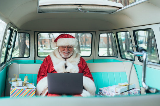 Cheerful man in costume of Santa Claus sitting in van with laptop and colorful gifts on sunny day