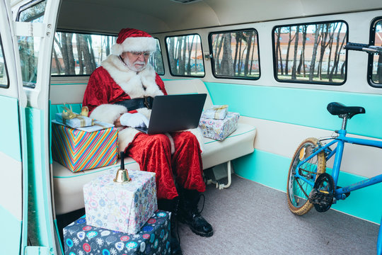 Cheerful man in costume of Santa Claus sitting in van with laptop and colorful gifts on sunny day